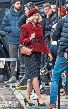 a woman in a red jacket and black skirt is walking down the street with other people behind her