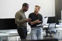 two men standing in an office talking to each other with computers on the desk behind them