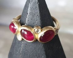 A gorgeous trio of rubies cabochons, the size of the stones are around 7x5mm, i have set them with 2 mm rose cut diamonds in the tones of cream and grays, beautifull combo, the entire ring is made of 18k yellow gold. Perfect to wear alone but can also be stack with your favorite bands. Made to order. Oval Ruby Cluster Ring With Rose Cut Diamonds, Heirloom Ruby Ring With Gemstone Accents, Three Stone Oval Ruby Ring, Cabochon Ruby Ring For Anniversary, Oval Rose Cut Diamond Ring With Ruby Accents, Heirloom Ruby Ring With Three Stones, Heirloom Ruby Three Stone Ring, Heirloom Ruby Three-stone Ring, Fine Jewelry Ruby Ring With Oval Cabochon