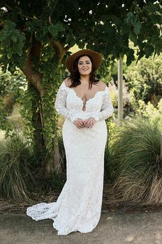 a woman in a white dress and hat standing under a tree with her hands on her hips