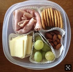 a plastic container filled with grapes, crackers and meats on top of a wooden table