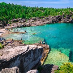 people are swimming in the clear blue water near some rocks and trees on the shore