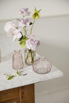 three glass vases with flowers in them on a marble top table next to a white wall