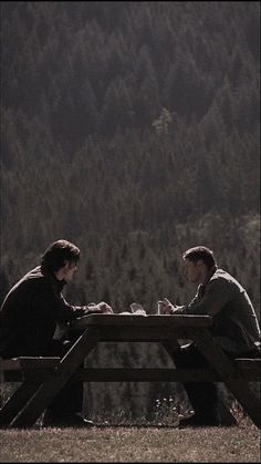 two people sitting at a picnic table in front of a forest with mountains behind them