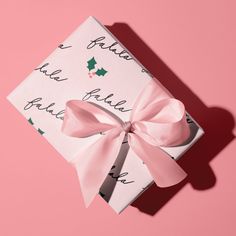 a present wrapped in white paper with pink ribbon and holly decorations on the top, against a pink background