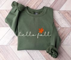 Celebrate your favorite season, Fall, with this moss olive green Hello Fall sweatshirt.  Welcome sweater weather and get cozy with warm apple cider in hand in this cute sweater. This sweater is made from polyester and cotton. This combination helps designs come out looking fresh and beautiful.  The collar is ribbed knit, so it retains its shape even after washing.   Knit in one piece using tubular knit, it reduces fabric waste and makes the garment more attractive.  There are no itchy side seams Khaki Cotton T-shirt For Fall, Khaki Cotton Tops For Fall, Fall Long Sleeve Khaki T-shirt, Khaki Long Sleeve T-shirt For Fall, Long Sleeve Khaki T-shirt For Fall, Khaki Crew Neck Tops For Fall, Fall Olive Crew Neck Top, Olive Crew Neck Top For Fall, Green Long Sleeve T-shirt For Fall