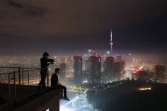 two people standing on top of a building looking at the city lights in the distance