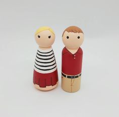 two wooden dolls are standing next to each other on a white surface, one is wearing a striped shirt and the other has a red dress