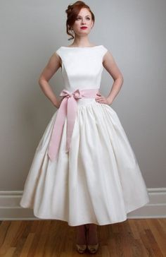 a woman wearing a white dress with a pink bow around the waist, standing in front of a gray wall