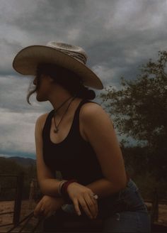 a woman wearing a cowboy hat sitting on top of a wooden fence next to a tree