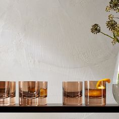 four shot glasses with an orange slice on the rim and one filled with ice, are lined up in front of a white wall