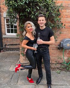 a man standing next to a woman holding a wine glass in front of a brick wall