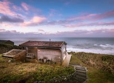 an old shack sitting on top of a grassy hill next to the ocean with stairs leading up to it