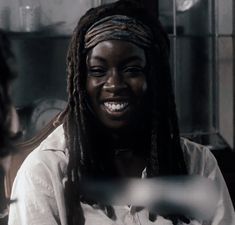 a smiling woman with dreadlocks sitting in front of a glass case and looking at the camera