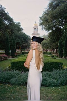 a woman wearing a graduation cap and gown standing in front of a garden with hedges