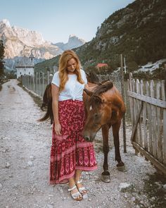 a woman standing next to a brown horse on a dirt road near a wooden fence