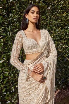 a woman standing in front of a bush wearing a white sari and matching jewelry