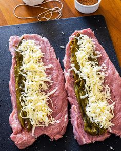 two hotdogs covered in cheese and pickles on a cutting board next to a bowl of seasoning