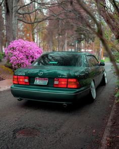 a green car parked on the side of a road next to some pink flowers and trees