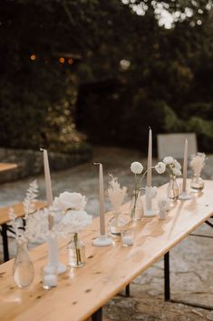 a long wooden table topped with vases filled with white flowers and candles next to each other