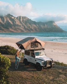 a woman standing next to a tent on top of a white truck near the ocean