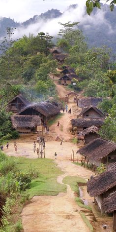 people are walking down a dirt road with huts on the sides and trees in the background