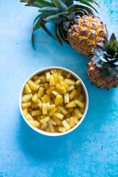 two pineapples are next to a small bowl of food on a blue surface