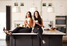 two women sitting on a kitchen counter with their legs crossed and holding coffee mugs