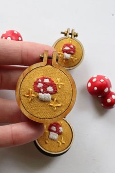 a hand is holding two small red and white mushroom brooches with gold thread on them