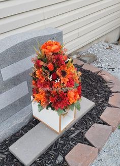 an arrangement of flowers in a white vase on a stone block near a wall and brick walkway