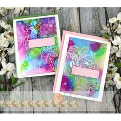 two cards with flowers on them sitting next to some white and pink flowers in the background