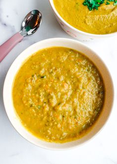 two bowls filled with soup on top of a white counter next to a spoon and pink utensil