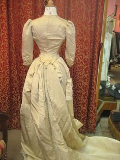 "1880's, 34\" bust, ecru silk faille, bustled and trained skirted wedding gown. A 19th century take on an 18th century lace up, low cut bodice. Bodice has a sharply pointed bottom, below waist, and 3/4 length cuffed sleeves. The bodice curves over hips, and is shorter in back than front. Bustled skirt has steel covered hoops built into back of skirt , and elaborate drapery forming into a long long train. Skirt has scalloped edge at bottom of the front, the bottom of train has a covered dust ruff Fitted Victorian Dress For Debutante Ball, Victorian Wedding Dress With Boned Bodice, Vintage Victorian Ball Gown With Boned Bodice, Fitted Historical Victorian Dress For Wedding, Historical Victorian Ball Gown With Fitted Bodice, Cream Victorian Dress For Costume, Beige Victorian Wedding Dress With Ruffles, Victorian Style Crinoline Ball Gown Dress, Fitted Cream Victorian Ball Gown