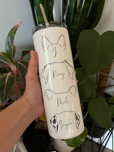 a hand holding a white tumbler with some drawings on it and plants in the background