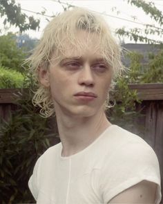 a man with blonde hair and white shirt looking at the camera while standing in front of a fence