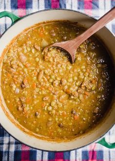 a pot filled with beans and meat on top of a checkered table cloth next to a wooden spoon