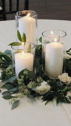 three white candles are sitting on a table with greenery and flowers around the candle holders