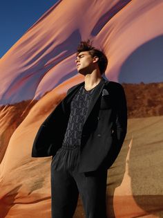 a man standing in front of a desert backdrop wearing a black jacket and pants with his hands on his hips