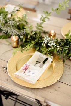 the table is set with gold plates and greenery, napkins and silverware