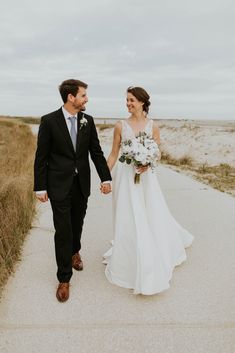 a bride and groom holding hands walking down a path in front of sand dunes at the beach