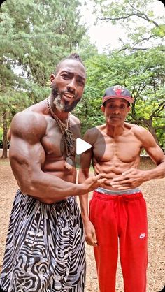 two men standing next to each other in front of trees and dirt ground with one man showing off his muscles