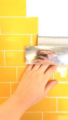 a hand is holding a tin foil over a yellow tiled wall with a mirror above it