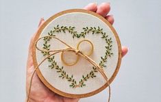 a hand holding a small embroidered heart with two gold wedding rings on it and green leaves around the hoop