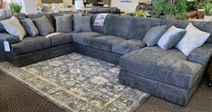 a large sectional couch with pillows on the floor in a furniture store display room, surrounded by rugs and other items