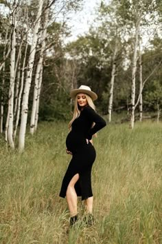 a pregnant woman in a black dress and hat standing in tall grass with her hands on her hips