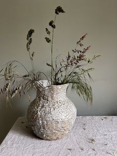 a vase with some plants in it sitting on a white table cloth next to a wall