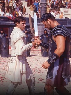 two men in roman costumes holding swords and facing each other with an audience behind them