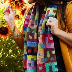 a woman wearing a scarf and holding a sunflower in her hand while standing next to some flowers