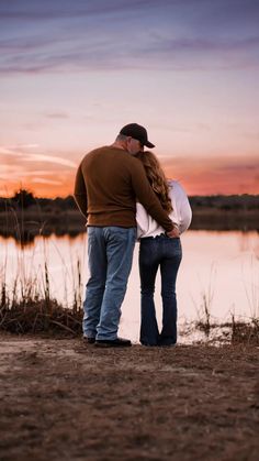 a man and woman standing next to each other on the shore of a lake at sunset