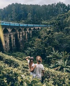 two people standing in front of a train going over a bridge